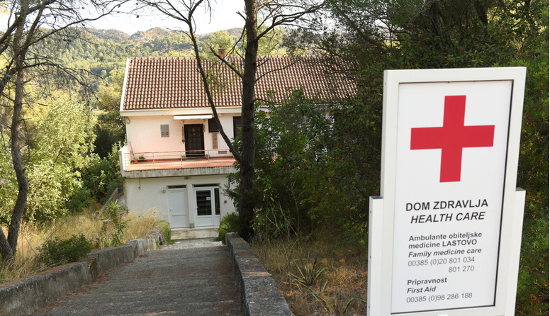 Vidi se dio zgrade okružen bujnim zelenilom. Do zgrade vodi šitoki put. u prvom planu je ploča na kojoj piše Dom zdravlja, na hrvatskom i engleskom jeziku.Ambulante obiteljske medicine Lastovo.Na tabli su i brojevi telefona..