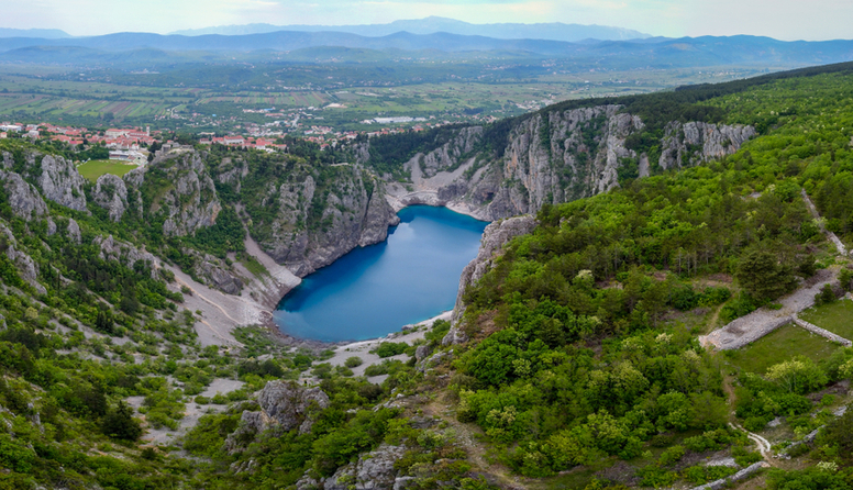 Fotografija iz ptičje perspektive prikazuje čisto, plavo jezero u obliku stopala, smješteno između krških brda i hridi, bijele boje, prošaranih zelenilom grmlja i krške vegetacije. U pozadini jezera i hridi naziru se svijetlo crveni krovovi kamenih kućica te nogometni stadion na uzvisini direktno iznad jezera.