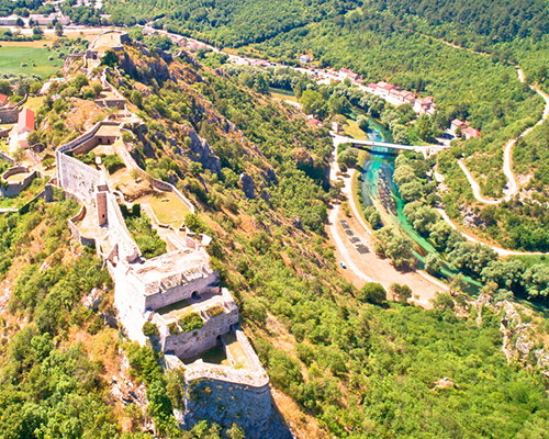 Planinski krajolik u podnožju kojeg je grad Knin kroz koji protječe rijeka Krka. Vidimo i most preko rijeke Krke i široku cestu uz grad.