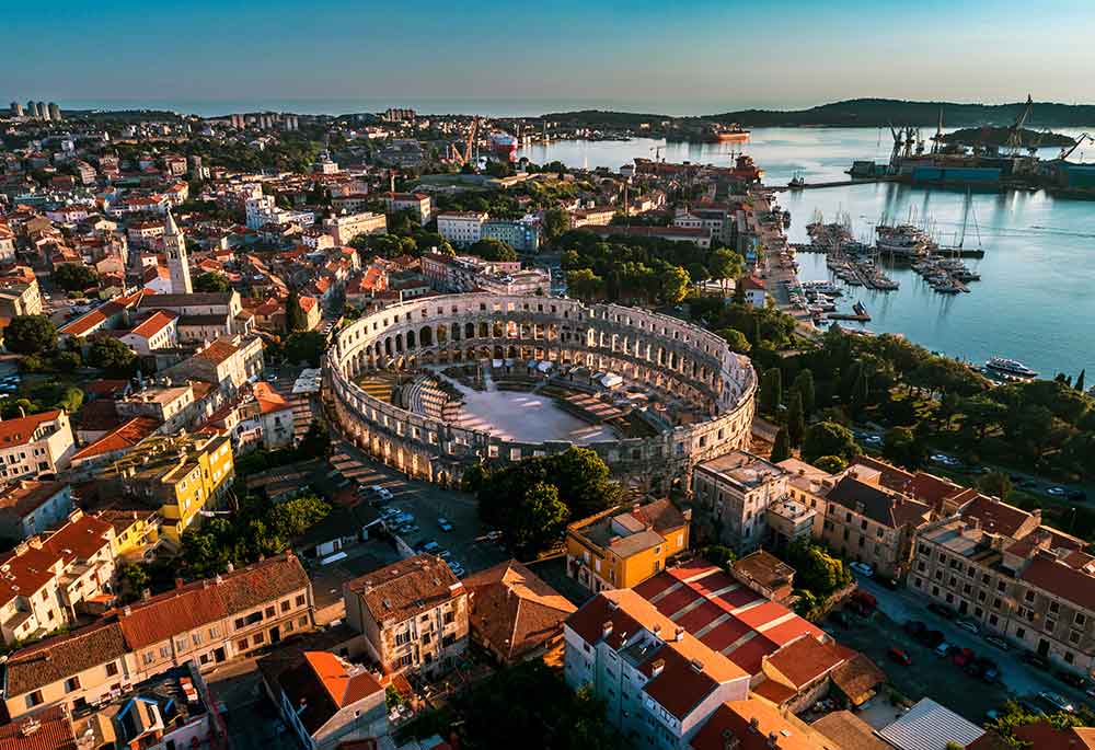 Panorama Pule s pogledom na Arenu. Do izražaja dolaze zidovi u obliku elipse s dvije razine lukova i gornjim dijelom s četvrtastim otvorima. U unutrašnjosti se vidi eliptični pano sa stepenastim gledalištem. Arena se nalazi u blizini obale.