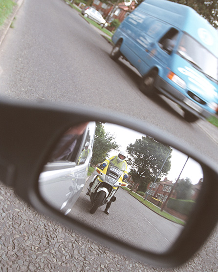 Prikazan je retrovizor automobila. U ogledalu retrovizora nalazi se policajac na motoru.