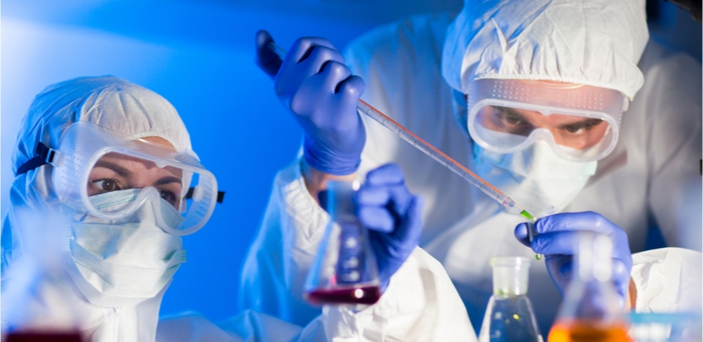 Fotografija prikazuje dvije osobe s punom zaštitnom opremom kako oprezno barataju kemikalijama u laboratoriju.