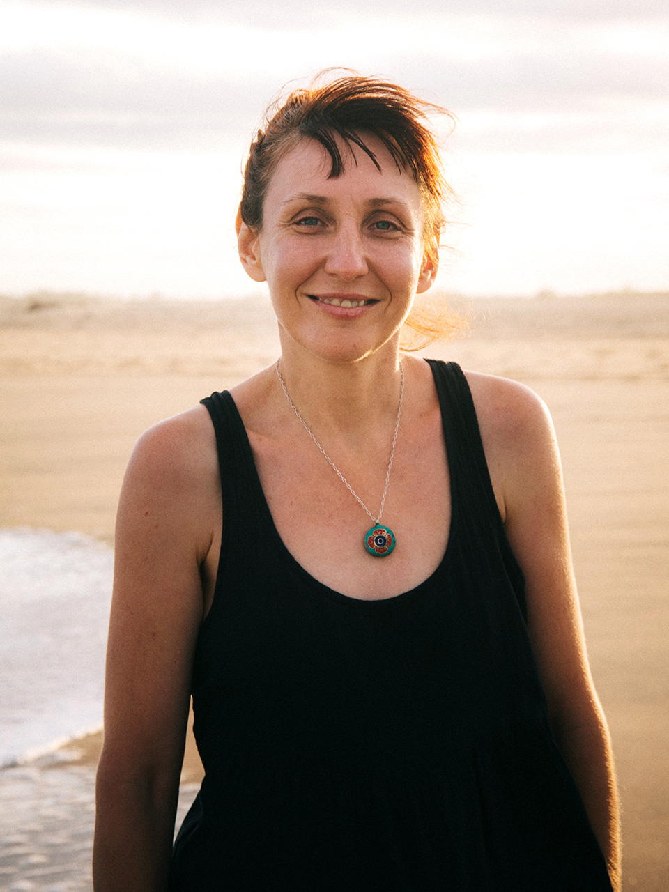 Žena srednjih godina u šetnji plažom kose svezane u rep i odjevena u crnu potkošulju. 