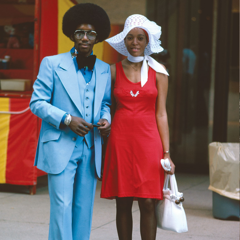 A man in a blue suit wearing an afro, a pair of sun glasses, a bow tie and a wrist watch & a woman in a red dress wearing wearing a white hat, a white scarf and a white bag.