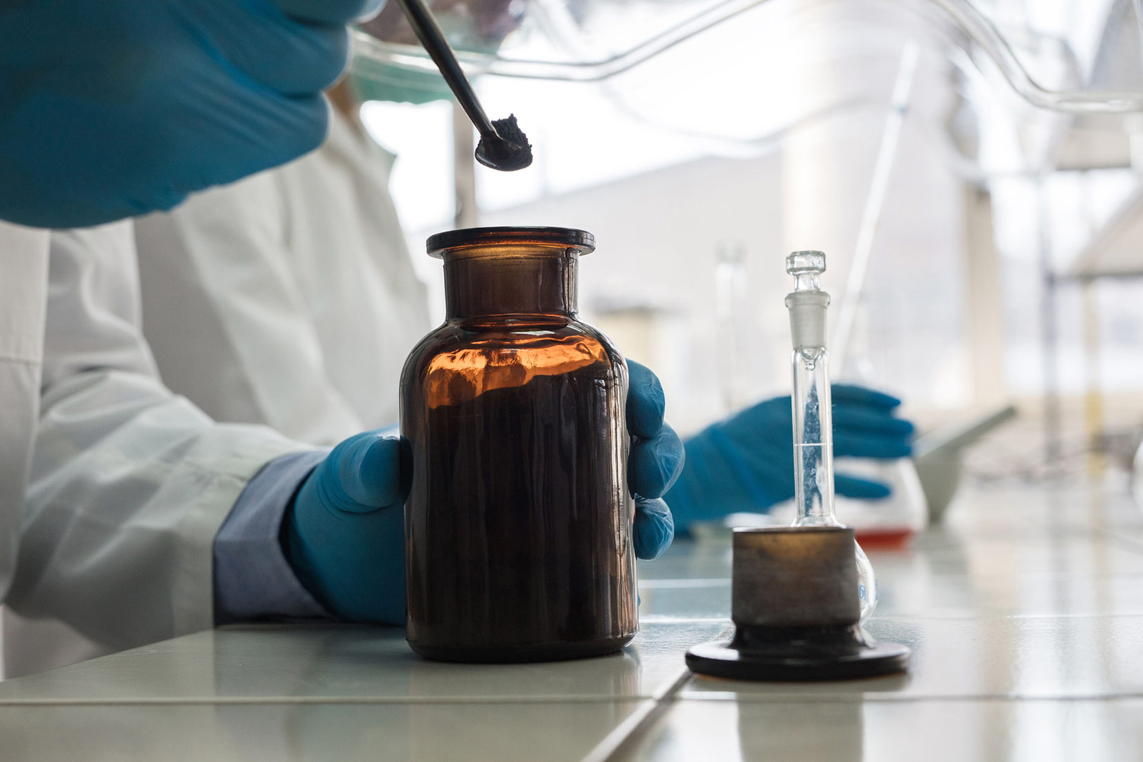Na slici je prikazan kemijski laboratorij i kemičareve ruke u plavim zaštitnim rukavicama . Iz bočice sa zatamnjenim staklom vadi uzorak (prah) sa žličicom.