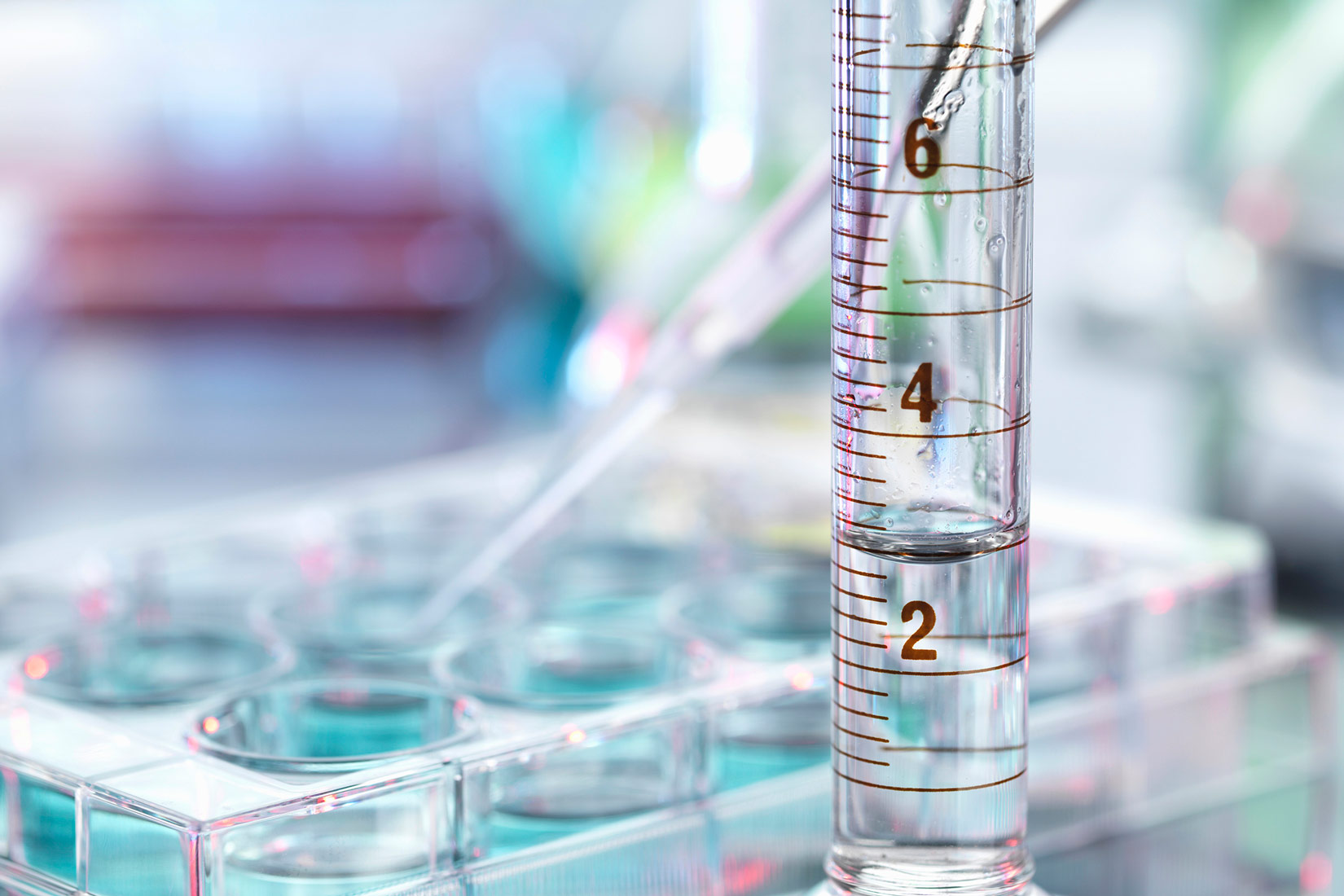 Na slici je prikazana menzura koja je graduirana (označene su crtice koje označavaju količinu tekućine). U njoj se nalazi prozirna tekućina.Iza se vidi staklena pipeta u obliku štapića.