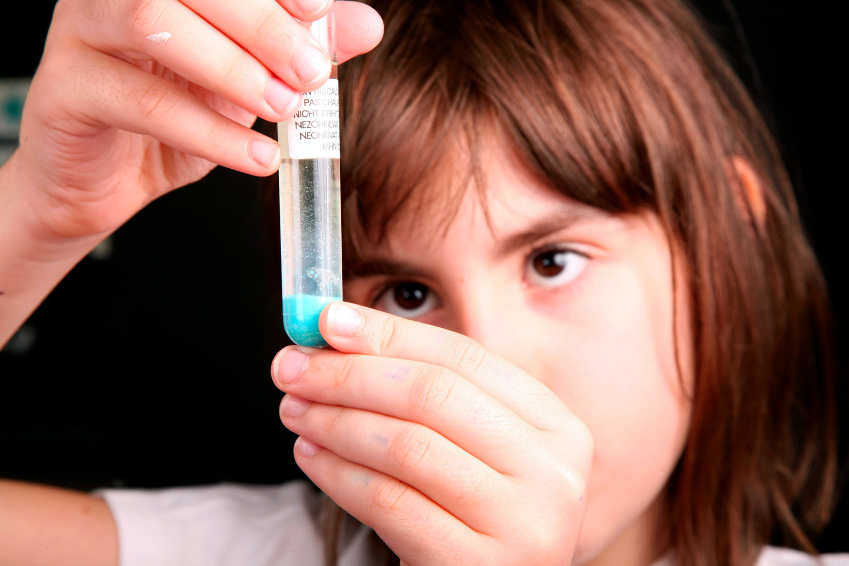 Na slici je prikazana djevojčica smeđe kose i očiju koja u rukama drži epruvetu s plavom tekućinom u visini očiju.