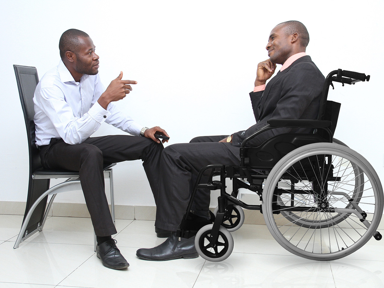 Dva formalno odjevena tamnoputa muškarca sjede jedan nasuprot drugom u opuštenom razgovoru. Dok jedan sjedi na stolici, drugi je u invalidskim kolicima.