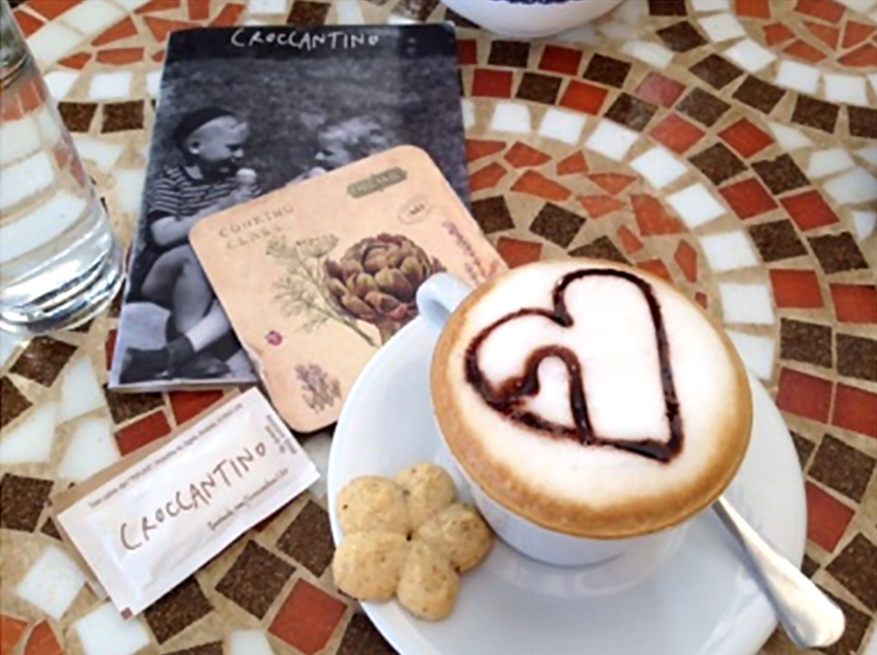 Na mramornom stolu kafića nalazi se šalica kave s puno pjene i ukrasnim srcem od čokolade dok je na tanjuriću keks u obliku cvijeta. 