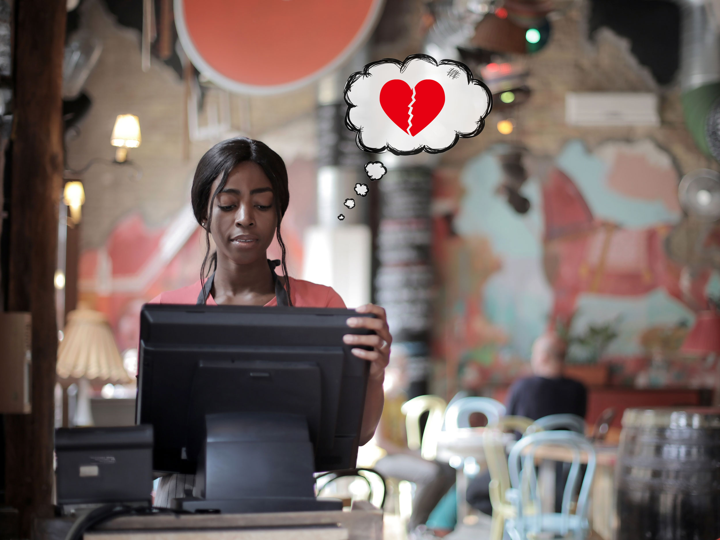 Tamnoputa djevojka stoji za blagajnom u restoranu i zamišljeno gleda u zaslon. U oblačiću pored njene glave je slomljeno crveno srce.