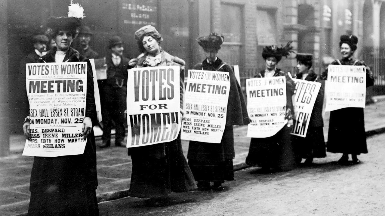Crno-bijela fotografija prikazuje šest sufražetkinja na gradskoj ulici kako nose plakate kojima se najavljuje sastanak na kojem će se tražiti pravo glasa za žene.
