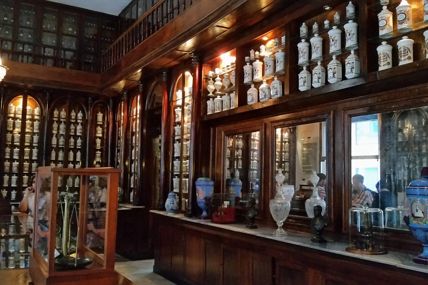 Fotografija prikazuje unutrašnjost ljekarne u kojoj na policama vidimo mnoštvo starih ljekarničkih posuda za čuvanje biljnog materijala. Posude su uglavnom bijele, a na njima su crteži i natpisi koji se odnose na biljni materijal koji se u njima čuva.