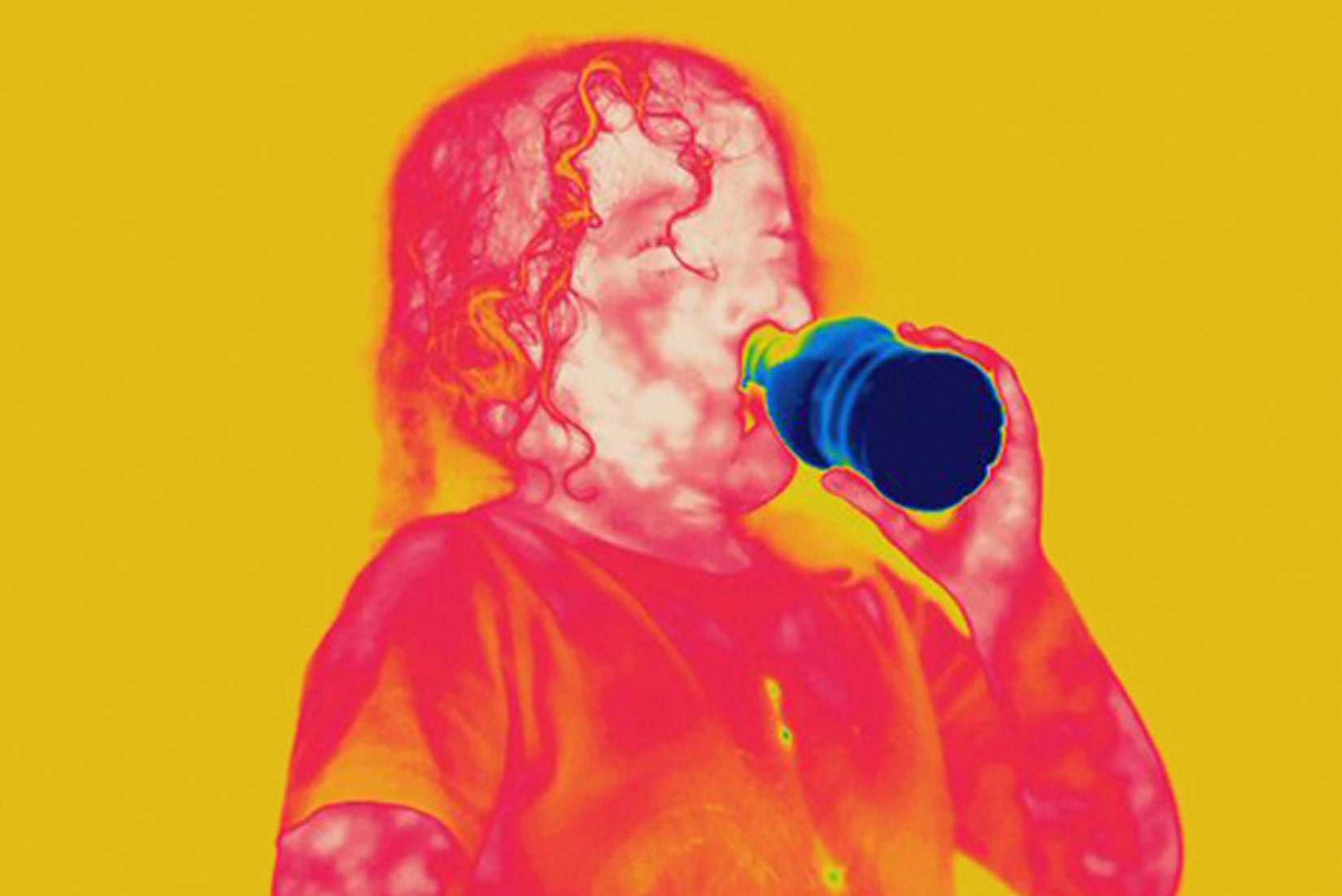 Slika 1. Prikazana je termografija, odnosno termalni snimak osobe koja pije piće. Osoba je obojana nijansama crvene boje, pozadina iza nje je žuta, a boca pića u njezinoj ruci je tamnoplave boje.