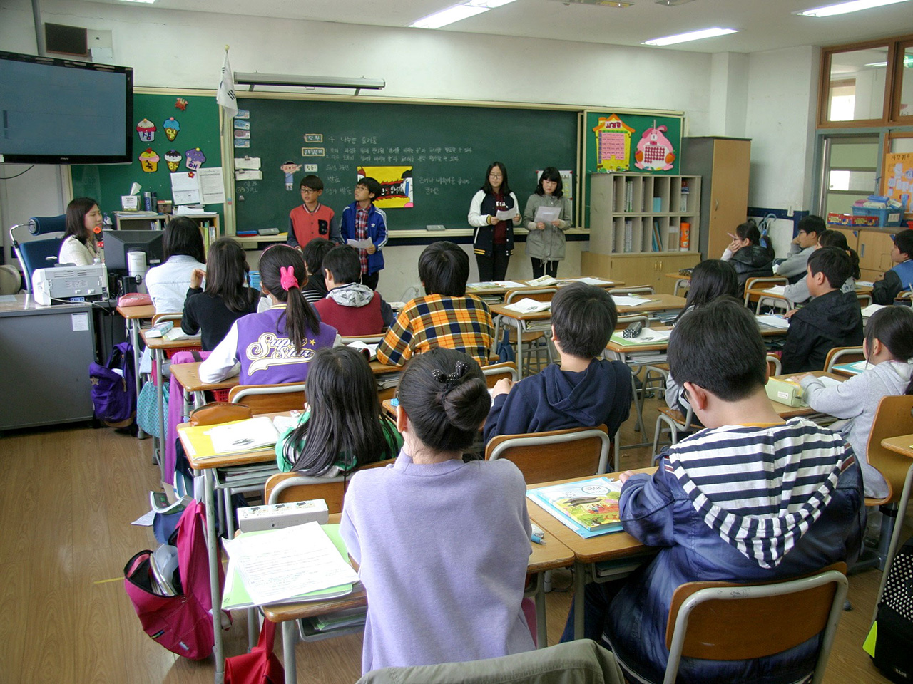 Učenici i nastavnica u učionici i nekoliko učenika pred pločom.