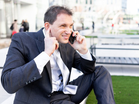 Muškarac sjedi na klupi u parku. Razgovara na mobitel na lijevo uho.  Kažiprstom lijeve ruke pokrio je desno uho.