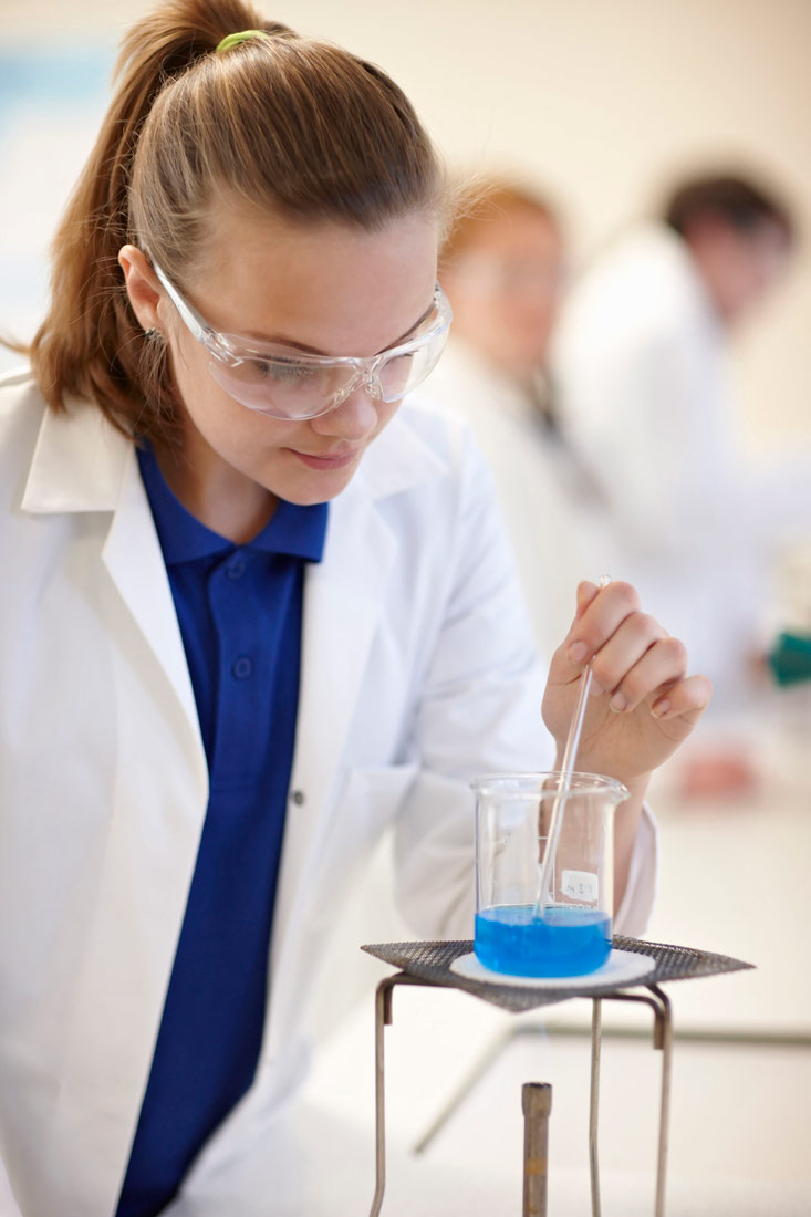 Slika prikazuje učenicu u laboratoriju koja miješa kemikaliju u staklenoj posudi iznad plamenika.Učenica ima zaštitne naočale.