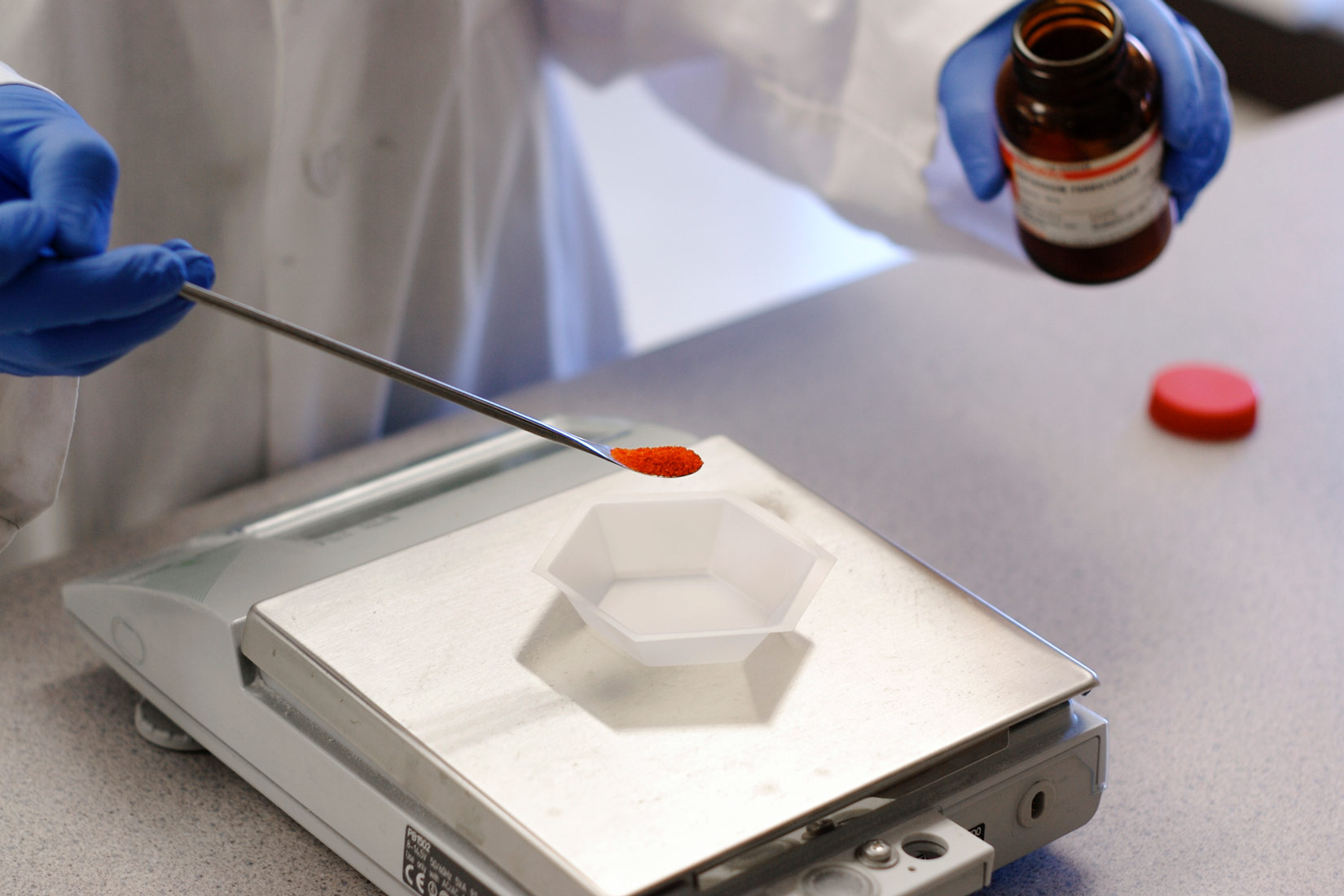 Na slici je prikazana digitalna laboratorijska vaga i ruke kemičara koji ima zaštitnu opremu (bijelu kutu, gumene plave rukavice).U desnoj ruci drži žličicu sa crvenim prahom a u lijevoj ruci drži tamnu otvorenu bocu.Na vagi se nalazi zdjelica bijela boje .