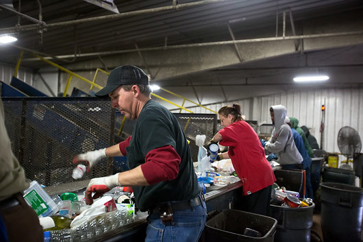 Fotografija prikazuje rad u pogonu za zbrinjavanje otpada. Radnici stoje pored trake na kojoj su različite vrte odpada te razvrstavaju otpad prema vrsti.