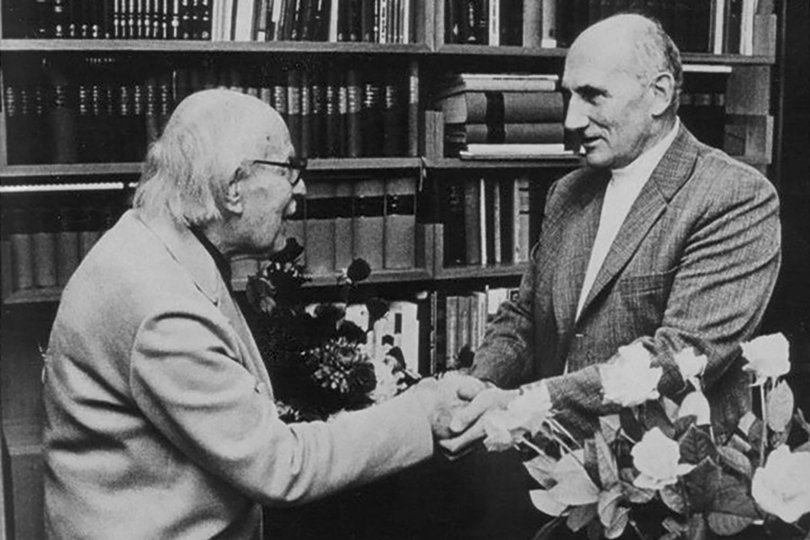 Fotografija prikazuje Lavoslava Ružičku i Vladimira Preloga, dobitnike Nobelove nagrade za kemiju. Rukuju se.U pozadini je polica s knjigama.U donjem desnom uglu se vidi cvijeće.