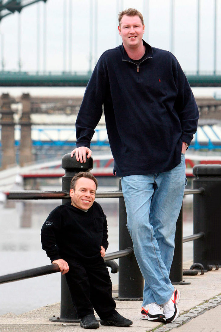 Fotografija prikazuje sliku dvojice muškaraca naslonjenih na ogradu mosta. Jedan je patuljastog rasta i niži je od ograde, a drugi je više nego dvostruko viši od njega.