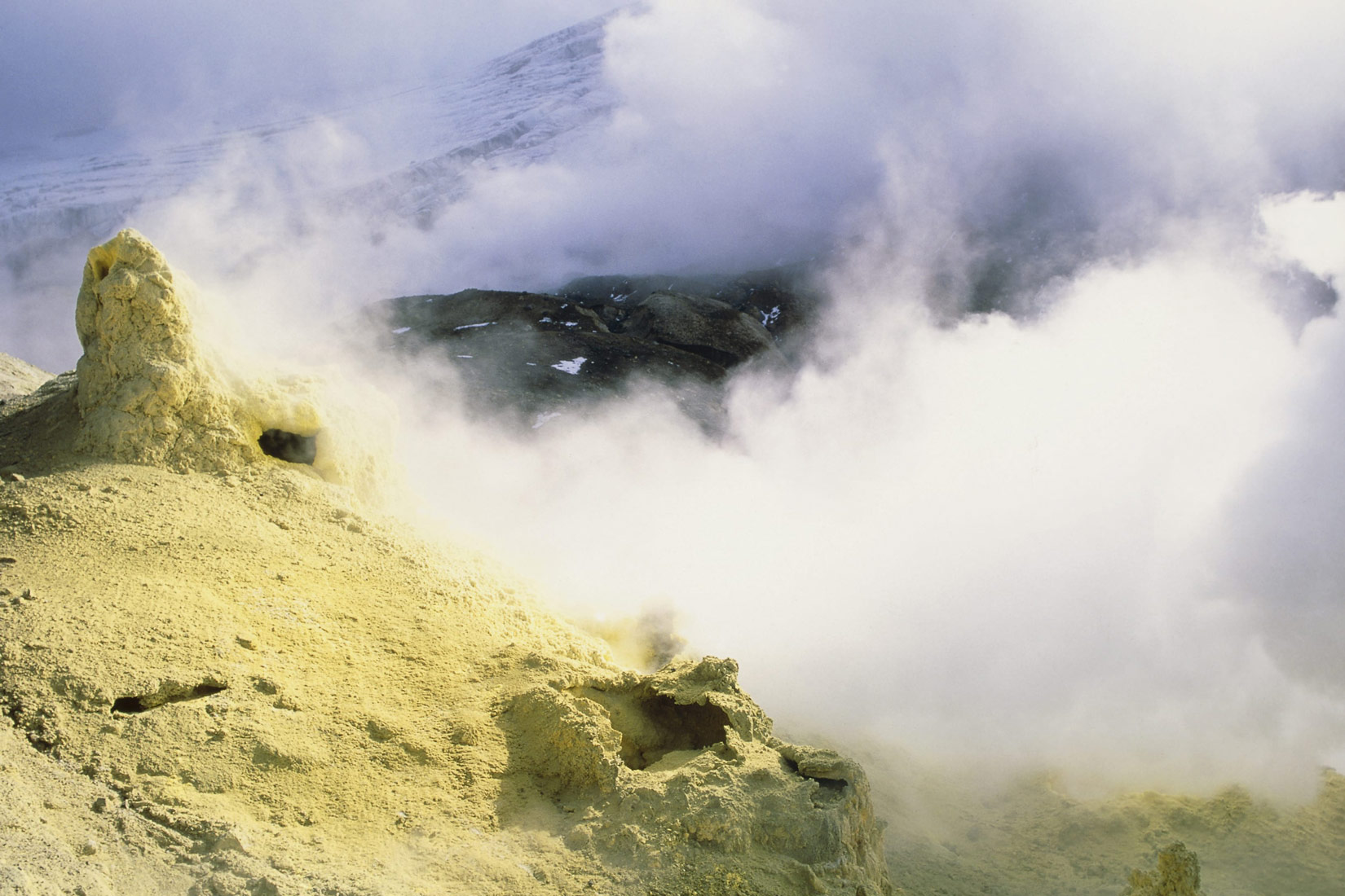 Fotografija prikazuje nalazište sumpora u prirodi. Vide se naslage sumpora koje izgledaju poput žutih stijena. Budući da su se naslage sumpora stvorile na vulkanskom krateru, na fotografiji vidimo i dim koji izlazi iz kratera.