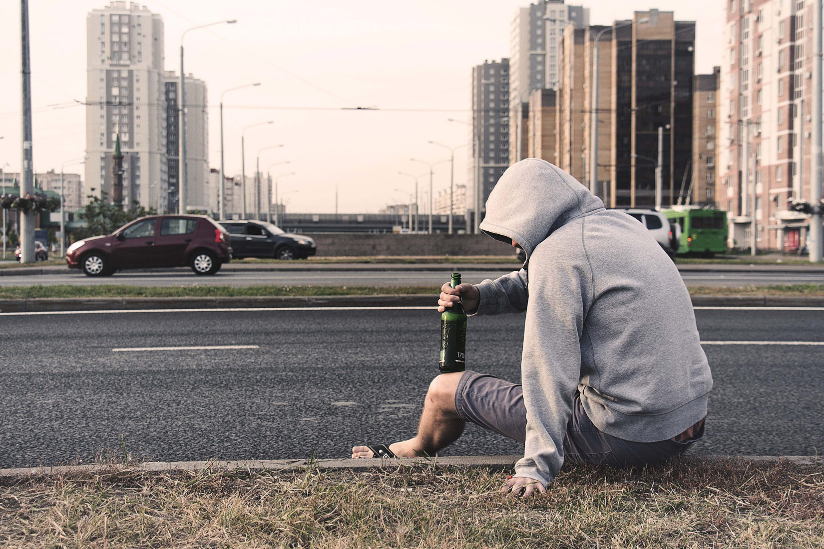 Fotografija prikazuje mladi čovjek ovisnog o alkoholu koji sjedi na rubu prometnice velikoga grada.