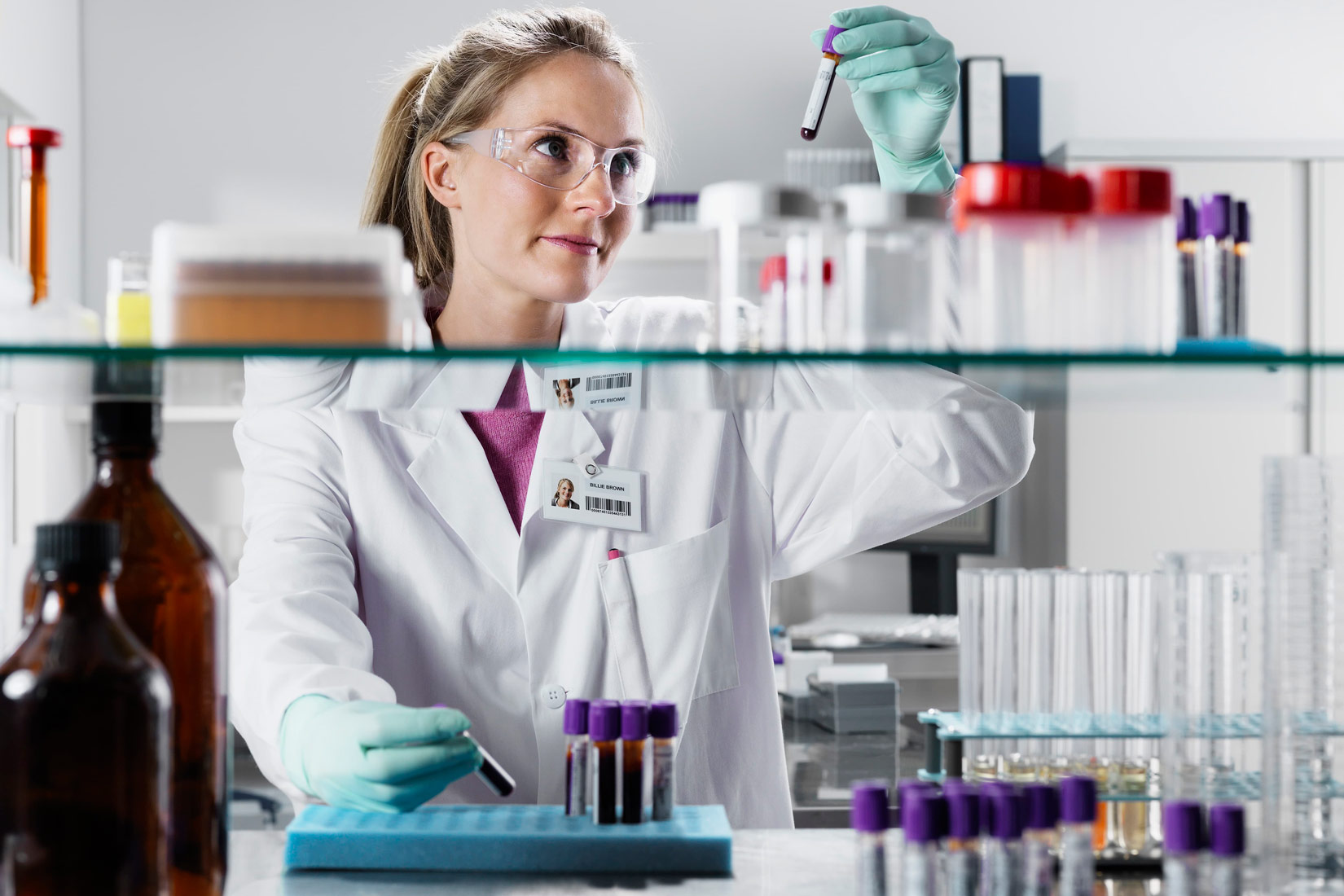 Na slici je prikazana kemičarka sa zaštitnom opremom (bijela kuta, zelene gumene rukavice) i skupljenom kosom. U ruci drži epruvetu s uzorkom. Ispred nje se nalazi staklena polica s kemijskim posuđem : lijevo dvije boce sa zatamnjenim staklom, desno stalak s epruvetama.