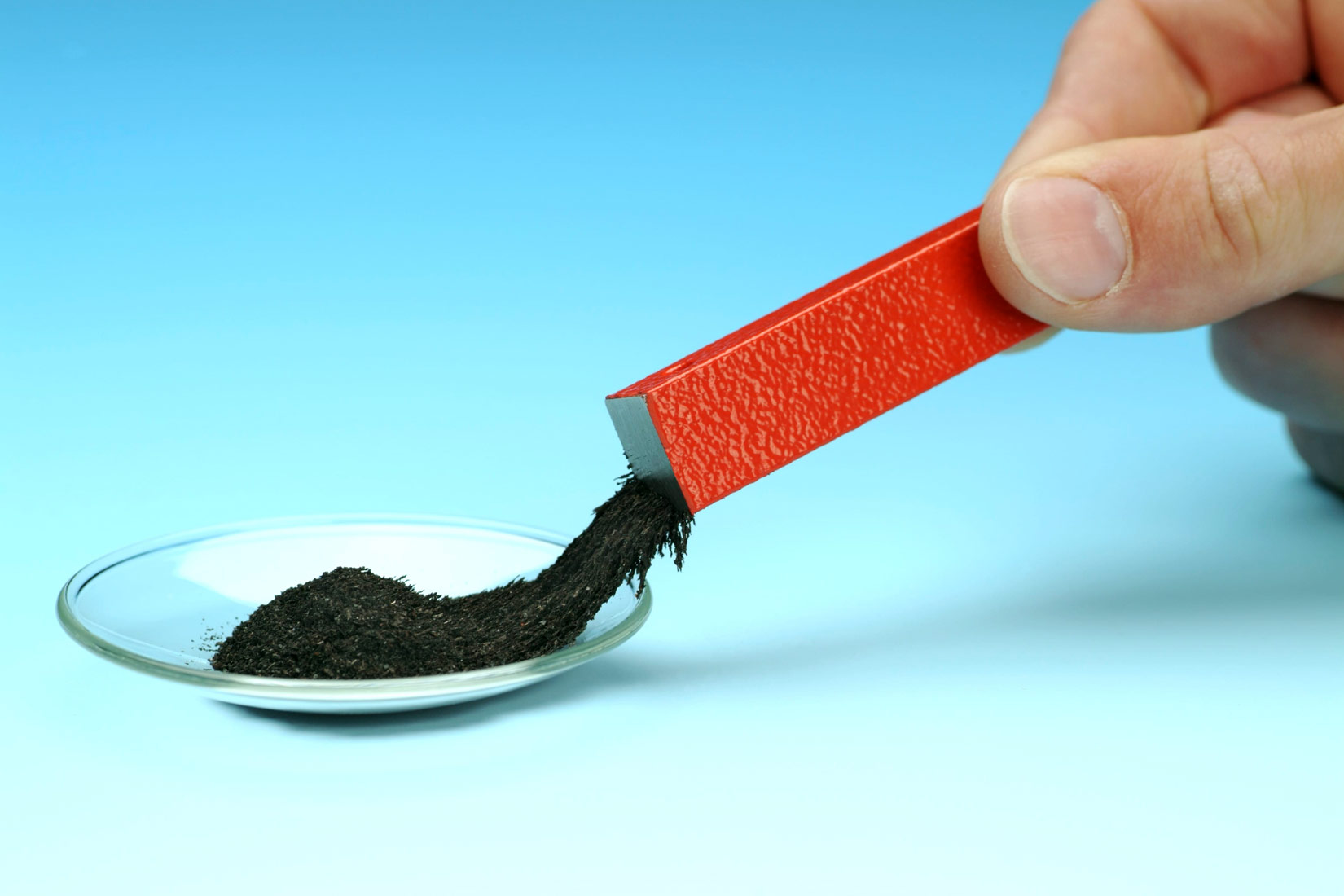 Na slici je prikazana ruka koja drži magnet crvene boje u obliku kvadra na plavoj podlozi. Strugotina željeza je crne boje i nalazi se u staklenom tanjuriću okruglog oblika. Magnet privlači željezo.