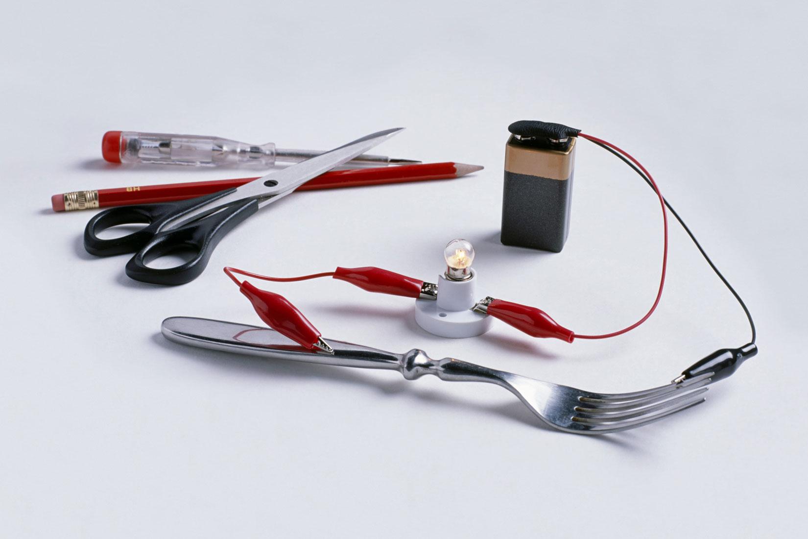 Fotografija prikazuje strujni krug koji se sastoji od vodiča, žarulje, baterije i vilice.Električna vodljivost je svojstvo tvari da vodi električnu energiju. U pozadini se još vide škare i grafitna olovka i odvijač koji može služiti kao ispitivač.