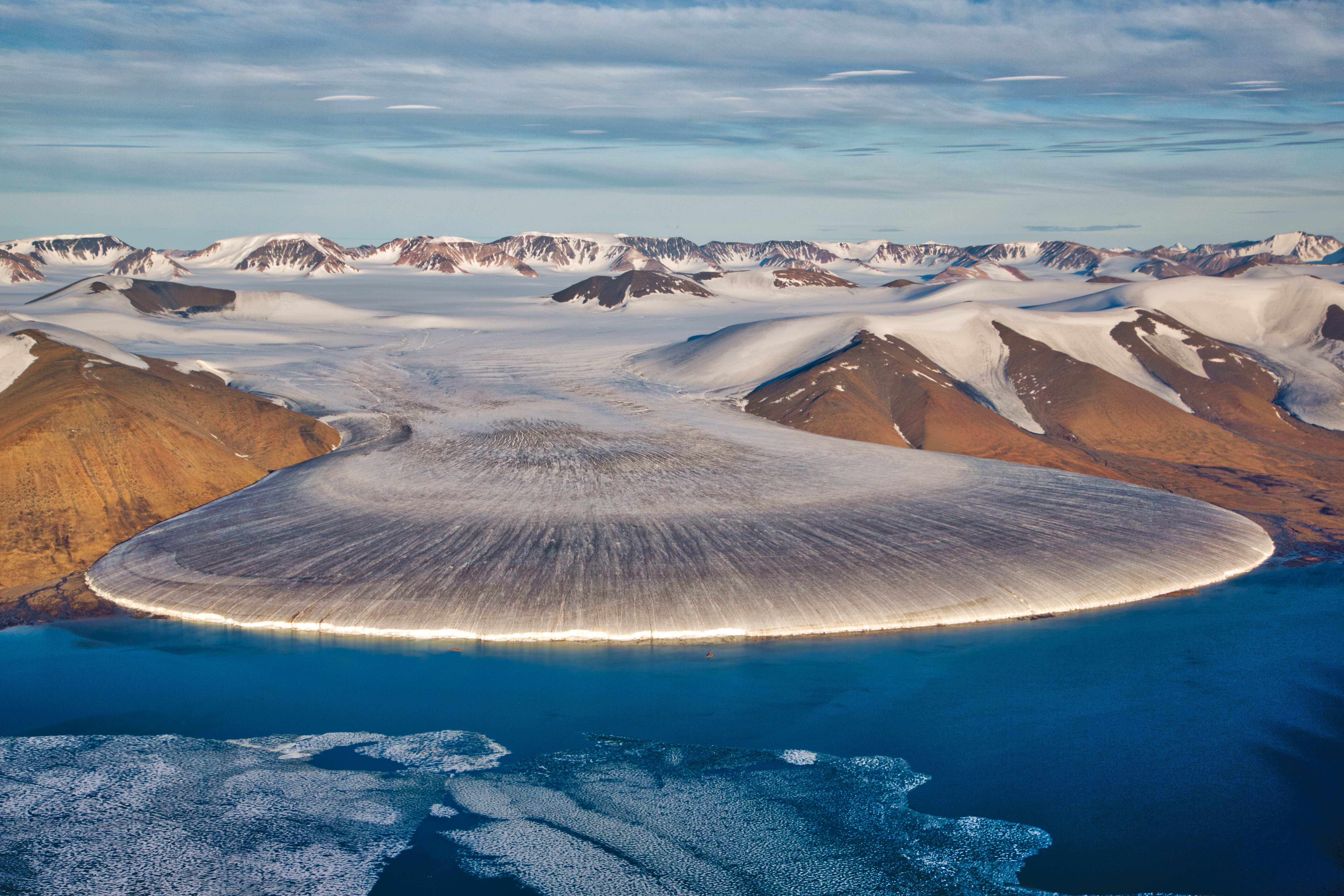 Ledenjak, Grenland. Fotografija prikazuje planinsko područje pod snijegom. Padine planina su smeđe boje i nisu prekrivene snijegom. Na mjestu dodira kopna i oceana nalazi se bijela ledenjačka masa u obliku školjke koja je nalik na tok lave koja utječe u more. 