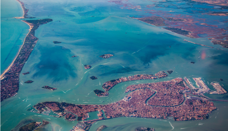 Venecija, Italija.  Fotografija prikazuje pogled iz zraka na grad Veneciju koja se smjestila u lagunu na nekoliko, što većih, što manjih otoka i otočića. Iz zraka je vidljivo mnoštvo crvenih krovova te mnogobrojni kanali