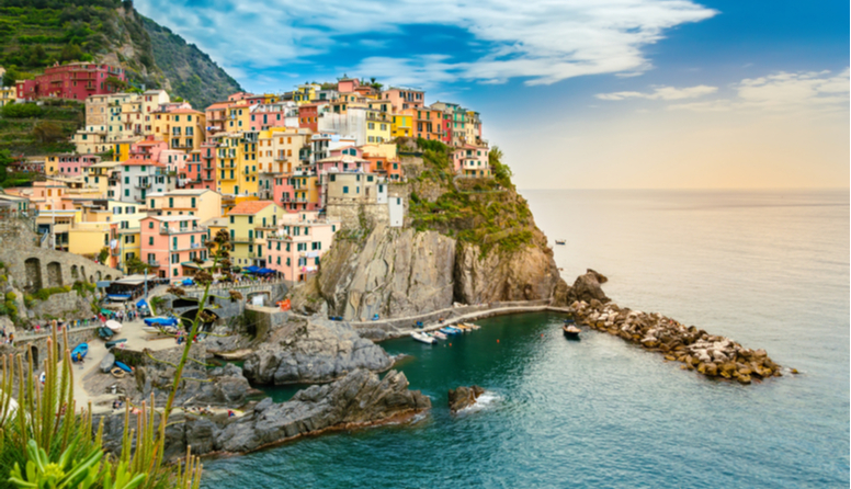 Cinque Terre, Italija. Fotografija prikazuje gradić na visokoj okomitoj hridi koja se izdiže iznaad mora. Gradić čine kućice šarenih fasada poput žute, roze, bijele, narančaste i crvene.