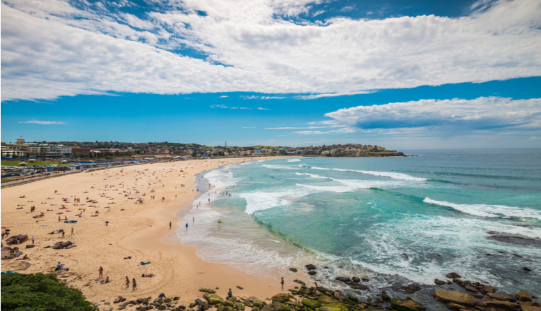 Plaža Bondi, Australija. Fotografija prikazuje široku i vrlo dugačku pješčanu plažu prekrivenu svijetlo smeđim pijeskom. Plažu oplakuje azurno more i valovi bijelih krijesti. Nebo je svijetle plave boje, na njemu se nalaze veliki, bijeli oblaci. Na plaži se sitni poput mrava uočavaju kupači