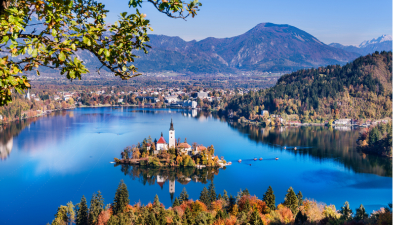 Jezero Bled. Fotografija prikazuje bistro, čisto jezero čije površina reflektira nebo, u čijem središtu se nalazi otočić kojim dominiraju kućice crvenih krovova i visoki crkveni toranj bijele boje i crnog krova