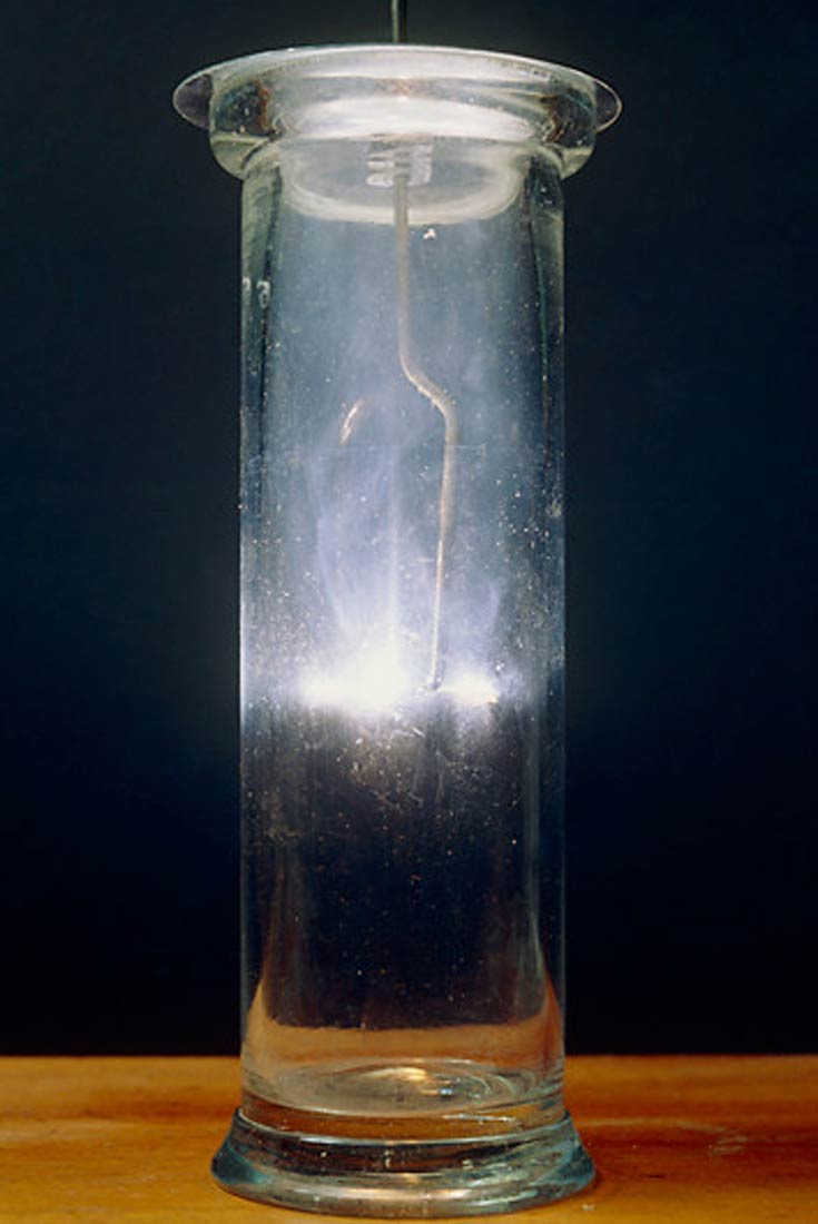 Fotografija prikazuje staklenu posudu valjkastoga oblika s poklopcem. U posudi je ovješen štapić s nanesenim kalcijem. Vidi se bjeličasto svjetlo kao posljedica gorenja kalcija u kisiku.