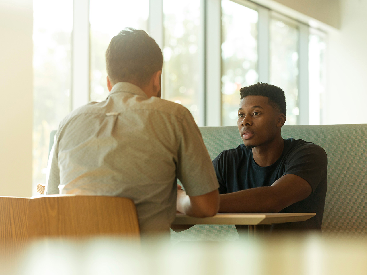 Dva muškarca sjede jedan nasuprot drugom za stolom i diskutiraju.