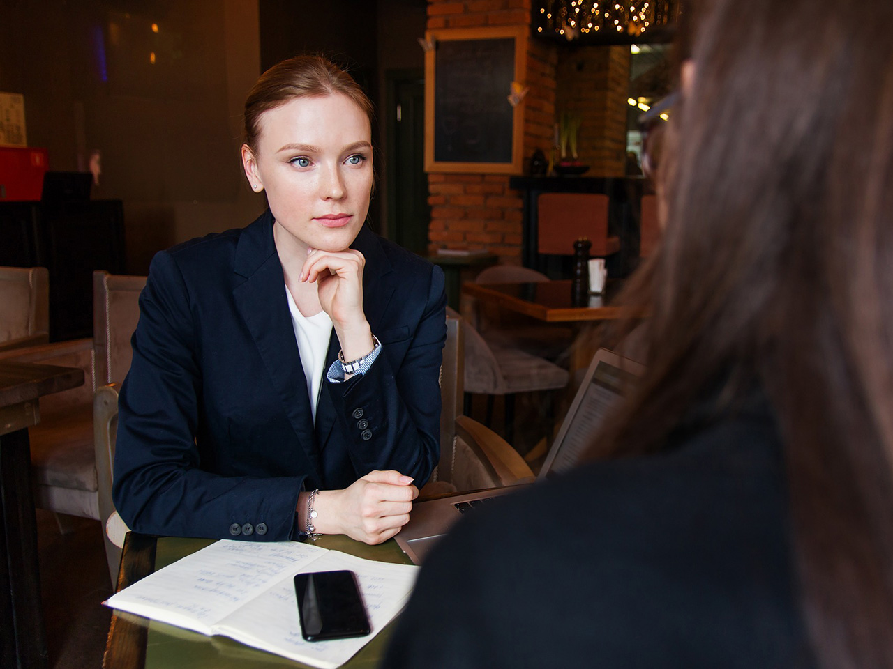 Žena u tamnoplavom kompletiću, brade naslonjene na prste lijeve ruke, pažljivo i ozbiljno promatra svoju sugovornicu. 