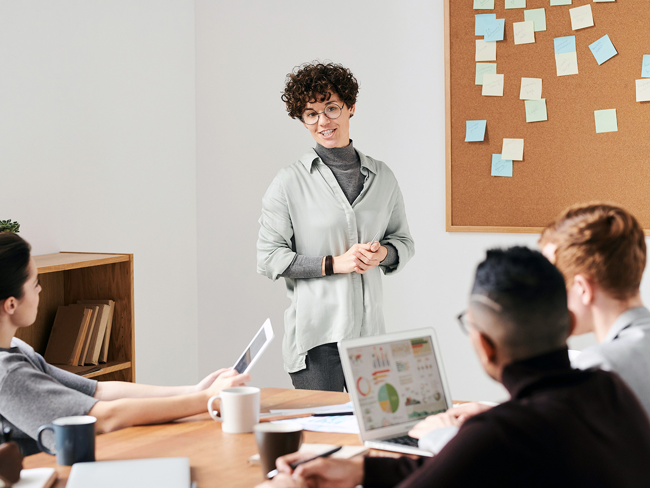 Žena kratke kovrčave kose i s naočalama stoji ispred plutene ploče pune post-it papirića i drži izlaganje kolegama s posla.