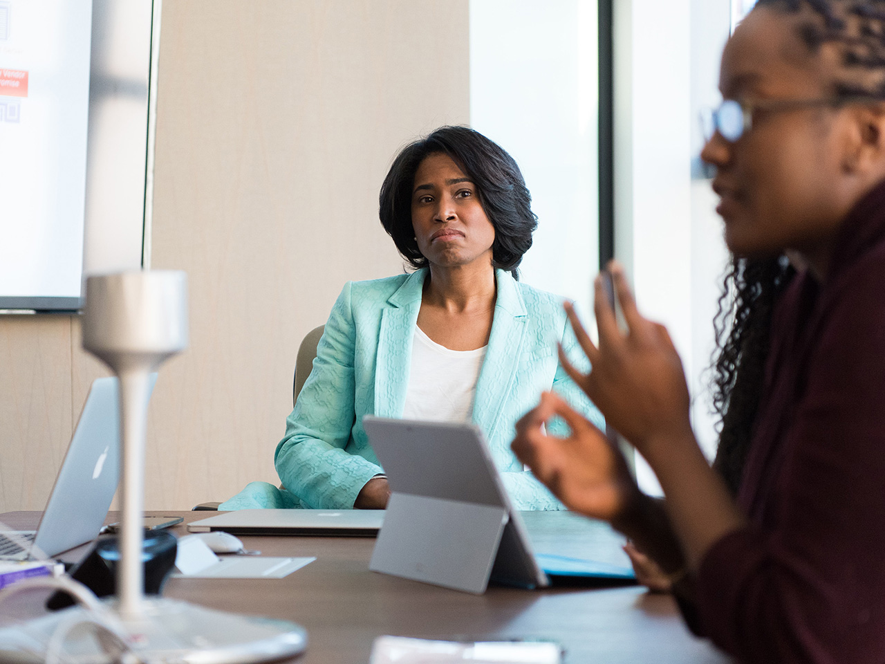 Tamnoputa žena u poslovnom kompletu sluša izlaganje kandidatkinje za posao i izraz lica otkriva da je sve samo ne oduševljena.
