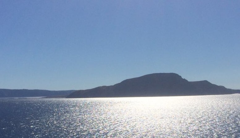 Goli otok. Fotografija prikazuje goli kameniti otok, okupan sunčevom svjetlošću pod vedrim, plavim nebom