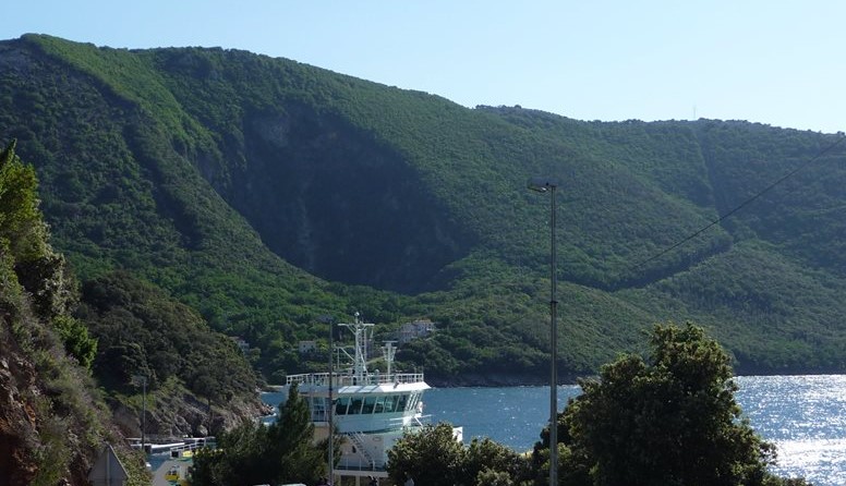 Trajektna luka Merag, otok Cres. Fotografija prikazuje malenu uvalu s lukom koja se smjestila u brdovitom krajoliku prekrivenom tamno-zelenom vegetacijom. U luci se nalazi usidren veliki bijeli trajekt