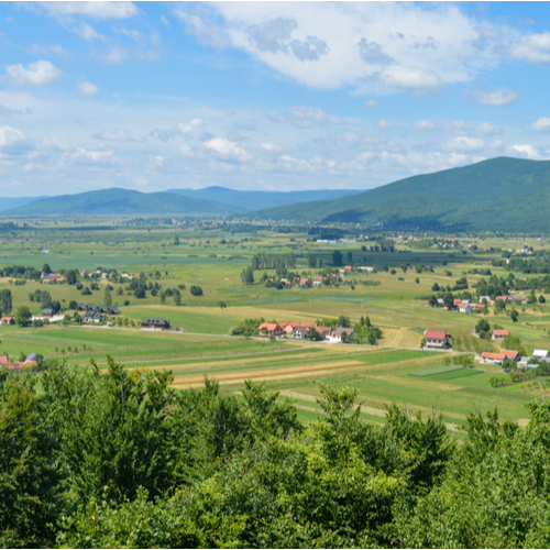 Polje u kršu. Fotografija prikazuje prostrano polje u daljini s raštrkanim kućama, a u daljini se vide obrisi uzvisina koje okružuju polje. 