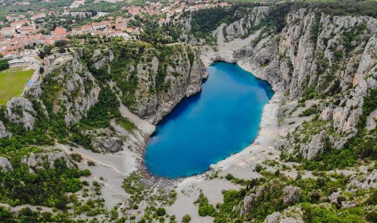 Fotografija iz ptičje perspektive prikazuje čisto, plavo jezero u obliku stopala smješteno između krških brda i hridi, bijele boje prošaranih zelenilom grmlja i krške vegetacije. U pozadini jezera i hridi naziru se svijetlo crveni krovovi kamenih kućica te nogometni stadion na uzvisini direktno iznad jezera.