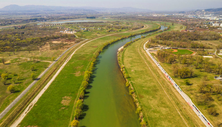 Široka mirna rijeka omeđena niskim grmljem, uz rijeku je široka livada s obje strane obale,pa nasip. Nakon nasipa s desme strane vidljive su visoke zgrade.