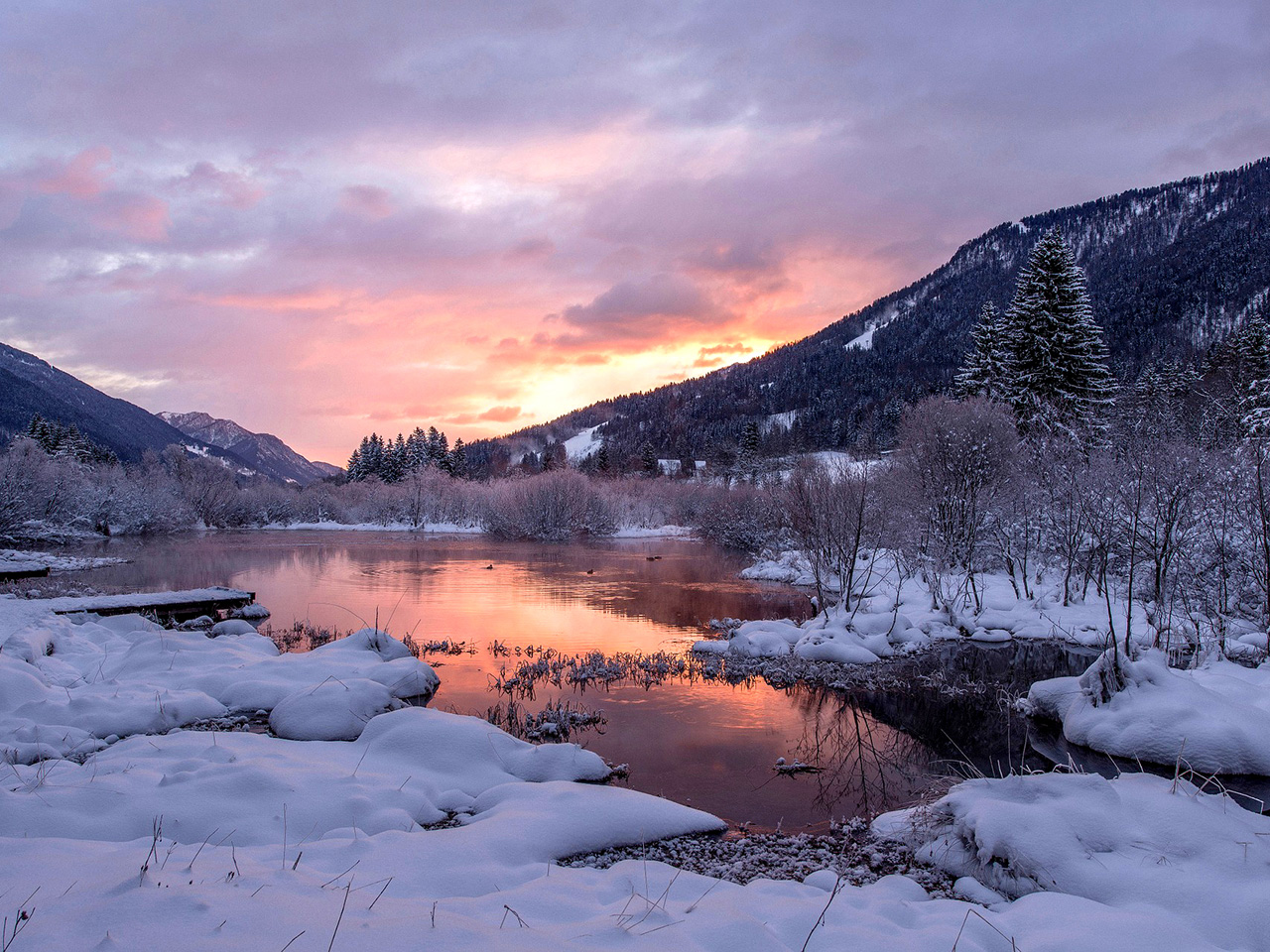 Zalazak sunca nad jezerom okruženom planinama i bogatom vegetacijom prekrivenom snijegom. 
