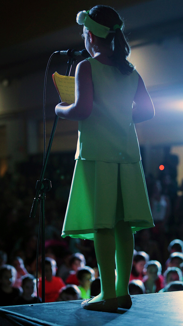 Djevojčica u zelenoj haljini stoji na pozornici i putem mikrofona se obraća drugoj djeci u publici. Također prizor s LiDraNa, državne smotre dramskog, literarnog i novinarskog dječjeg stvaralaštva.