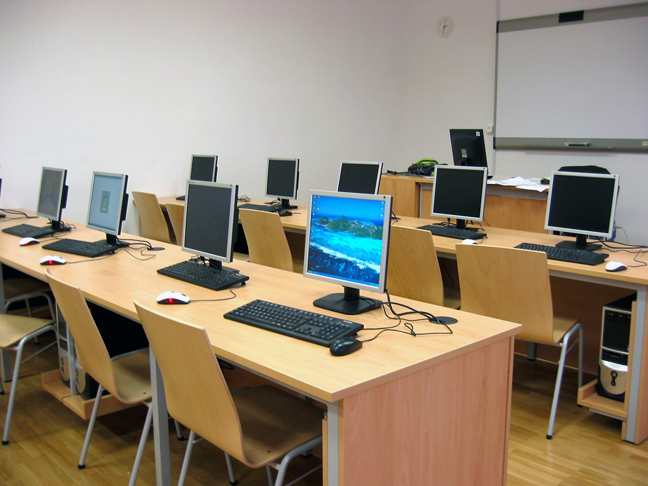 Informatička učionica s nizom klupa na kojima se nalaze monitori s tipkovnicama i miševima. U dnu učionice je katedra s računalom i bijela ploča. 