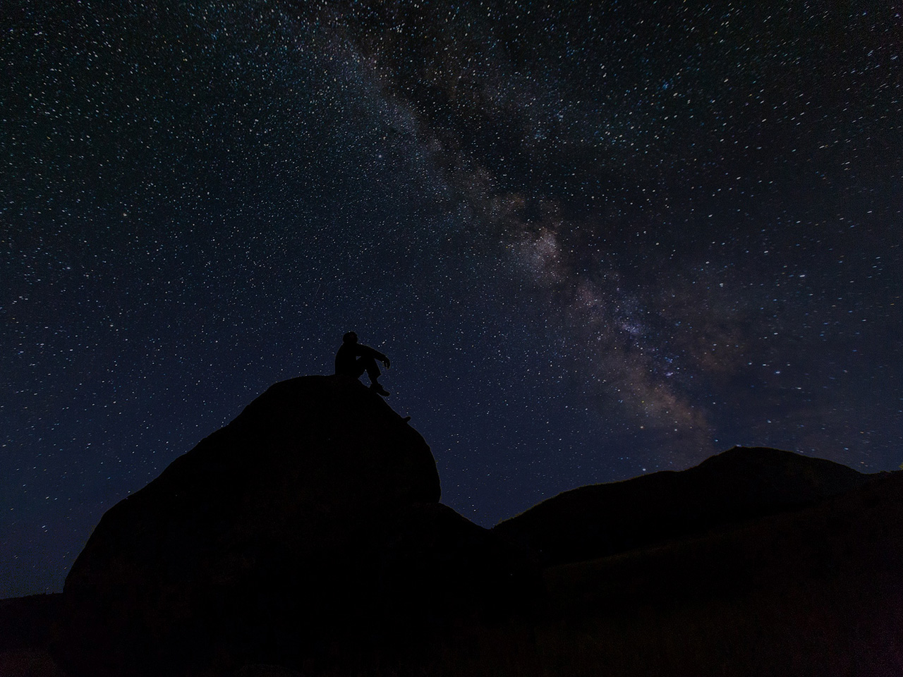Obris čovjeka koji sjedi na brdu i promatra noćno nebo s nebrojenim zvijezdama.