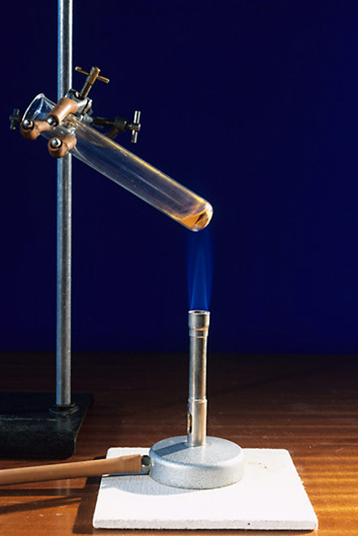 Fotografija krikazuje termolizu živinog oksida. Epruveta s a živinim (II) oksidom je pričvršćena za metalni stativ. Ispod je plamenik.Epruveta se zagrijava.Plamen je plave boje.