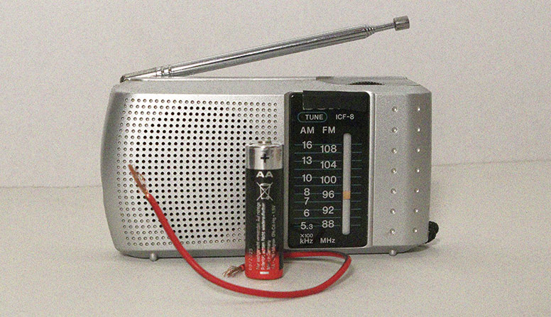 Na slici se nalazi:  prijenosni radio  s vanjskom antenom, sive boje, s desne strane su označene frekvencije, a s lijeve strane je zvučnik radija. Prikazana je i baterija od 1,5 V i  komadić crvene žice